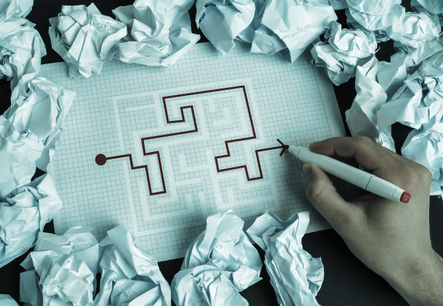Hand drawing a path through a maze on paper surrounded by crumpled sheets