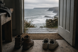 Two pairs of worn shoes by an open door, with a calm, grey coastal landscape beyond.